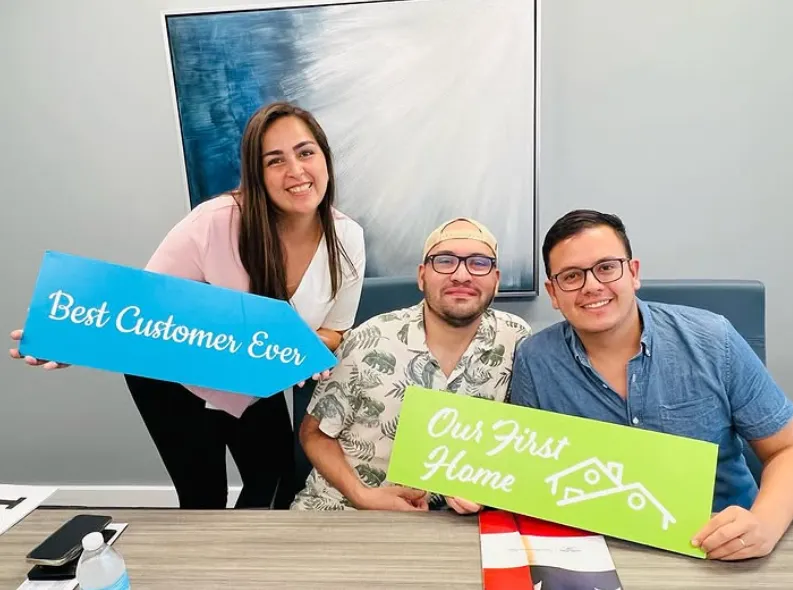 Gabriela with clients holding the "Best Customer Ever" and "Our First Home" signs on closing day